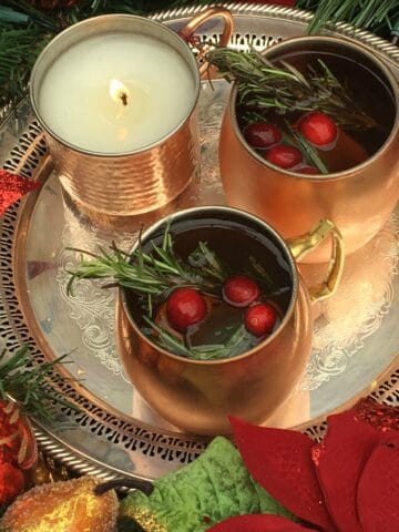 A fancy silver tray with a burning candle and two gold mugs of cocktail garnished with rosemary and cranberries