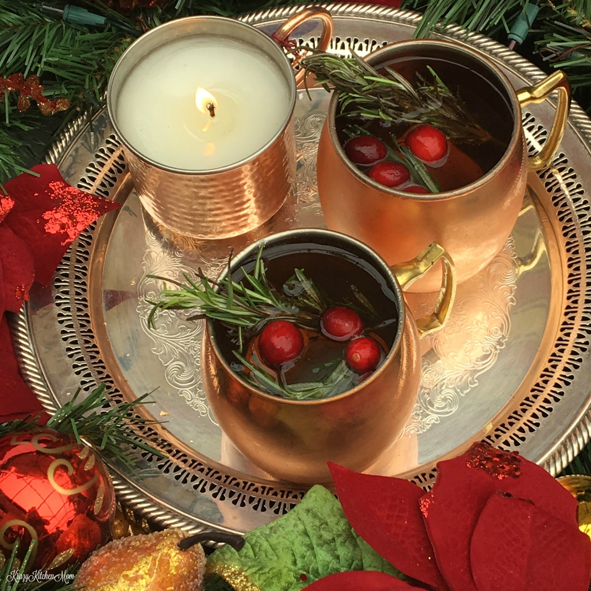 A fancy silver tray with a burning candle and two gold mugs of cocktail garnished with rosemary and cranberries