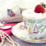 A tea cup filled with pink pudding and topped with strawberries with pearl on side and more tea cups in background