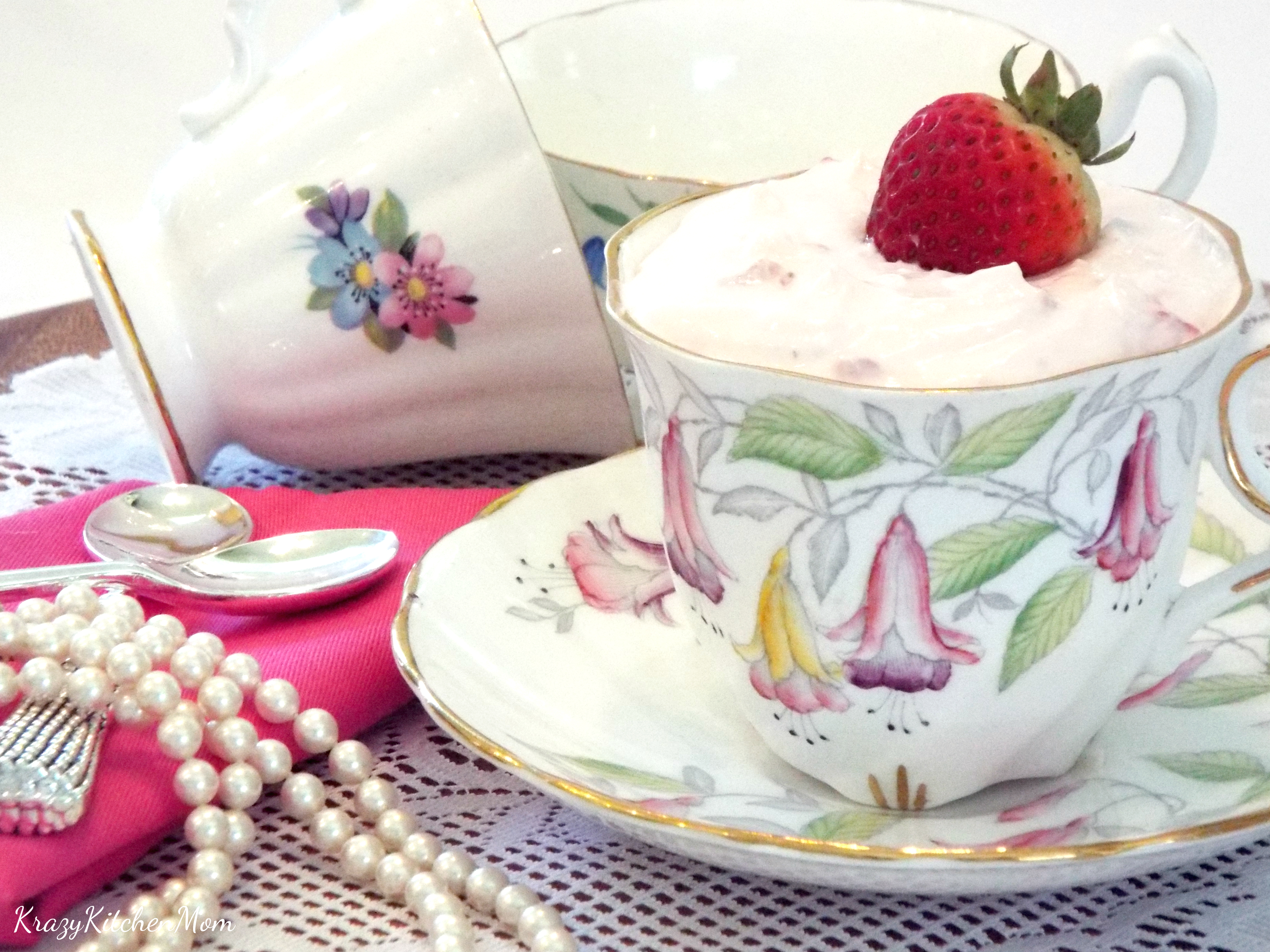 A tea cup filled with pink pudding and topped with strawberries with pearl on side and more tea cups in background
