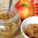 a jar of apples sauce with a spoon and a small dish and apple on the side