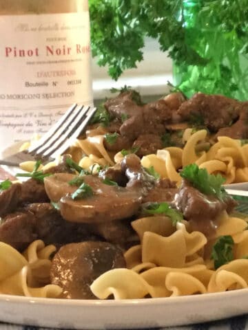 A plate of beef, mushrooms, noodles with wine bottle in background