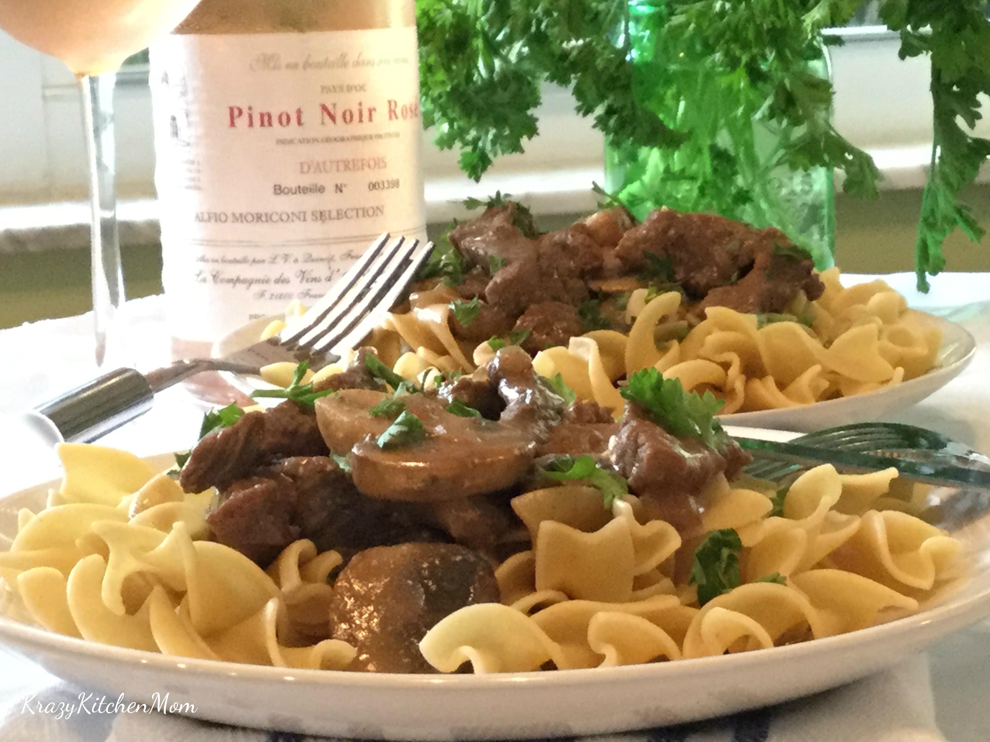 A plate of beef, mushrooms, noodles with wine bottle in background