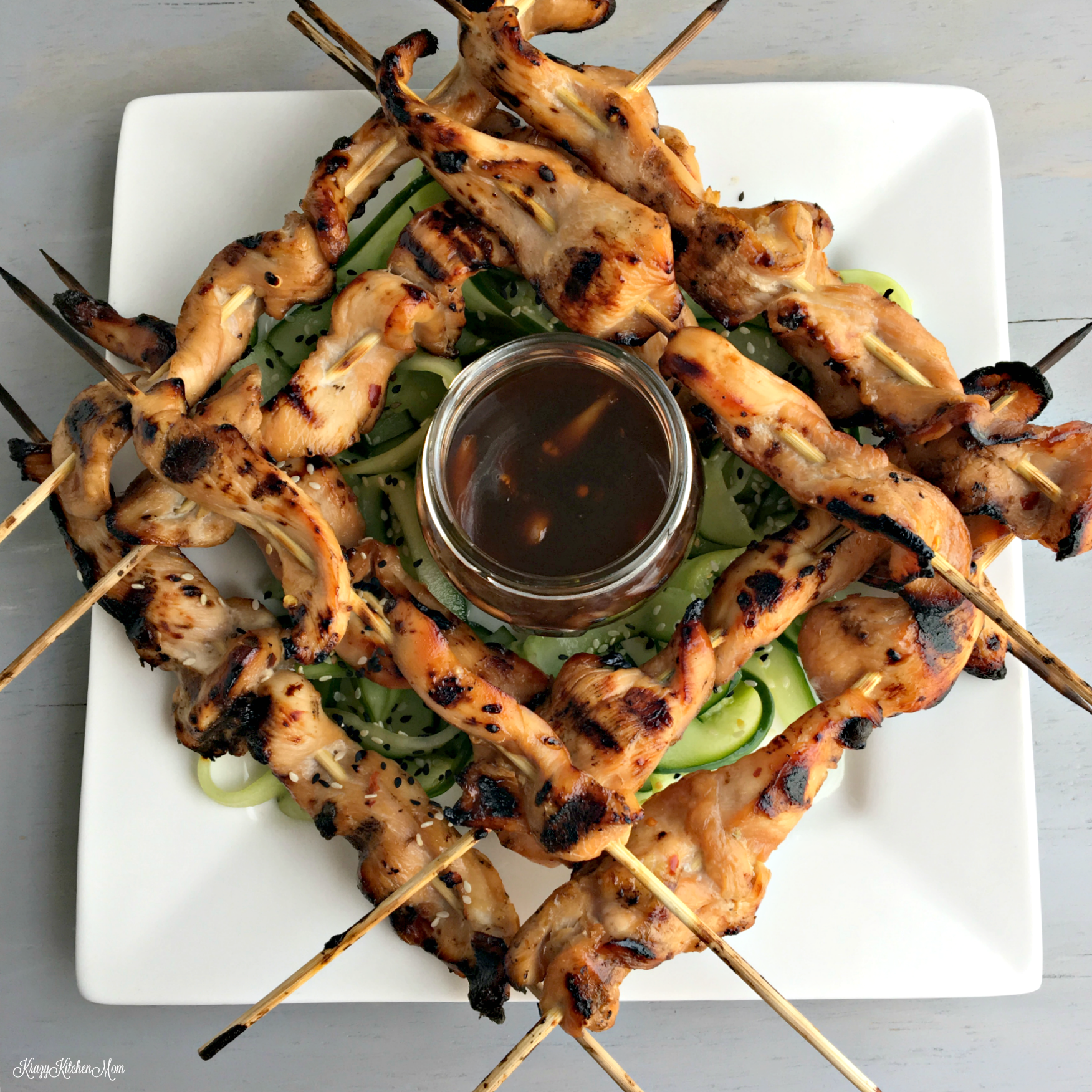 A plate with a crisscross of kabobs and a jar of sauce in the middle on a square white plate