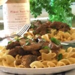 A plate of beef, mushrooms, noodles with wine bottle in background