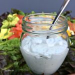 a close up of a jar of white salad dressing with veggies in background