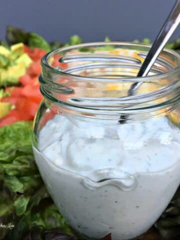 a close up of a jar of white salad dressing with veggies in background