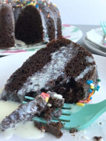 A slice of chocolate cake with white icing and a green fork with full cake in background