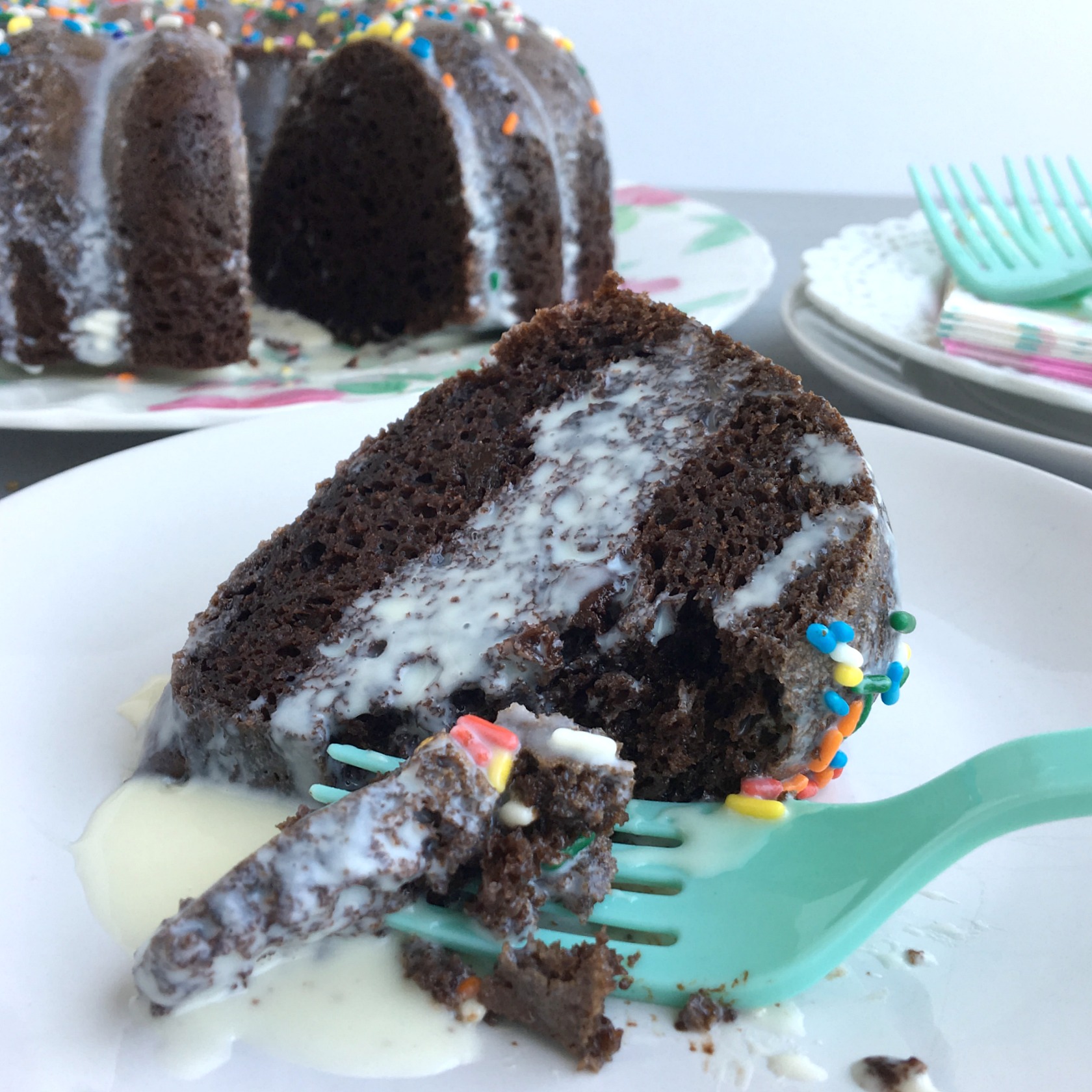 A slice of chocolate cake with white icing and a green fork with full cake in background