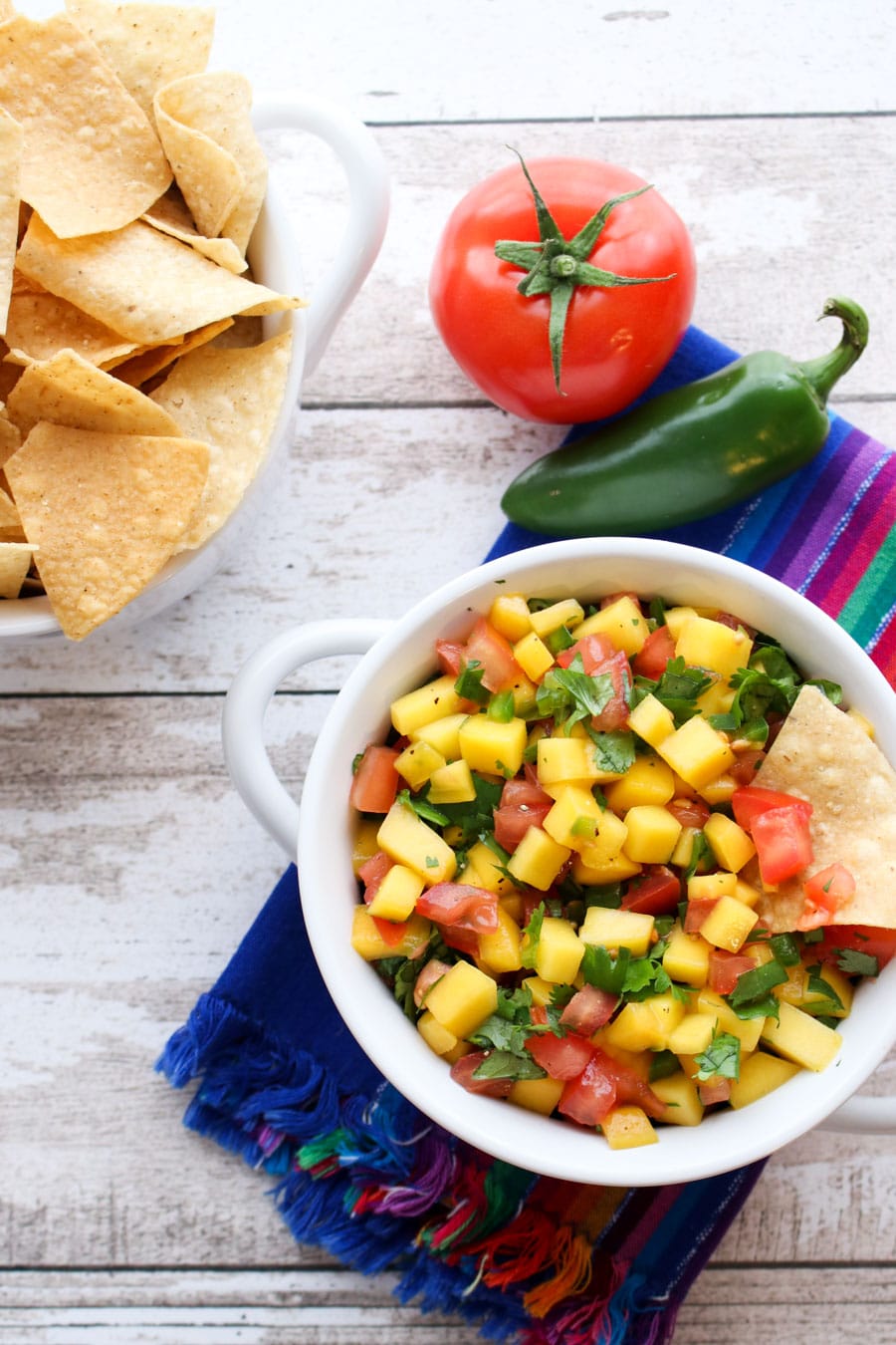 Bowl of mango salsa sitting on a blue colorful towel next to a dish of chips a tomato and a jalapeño.