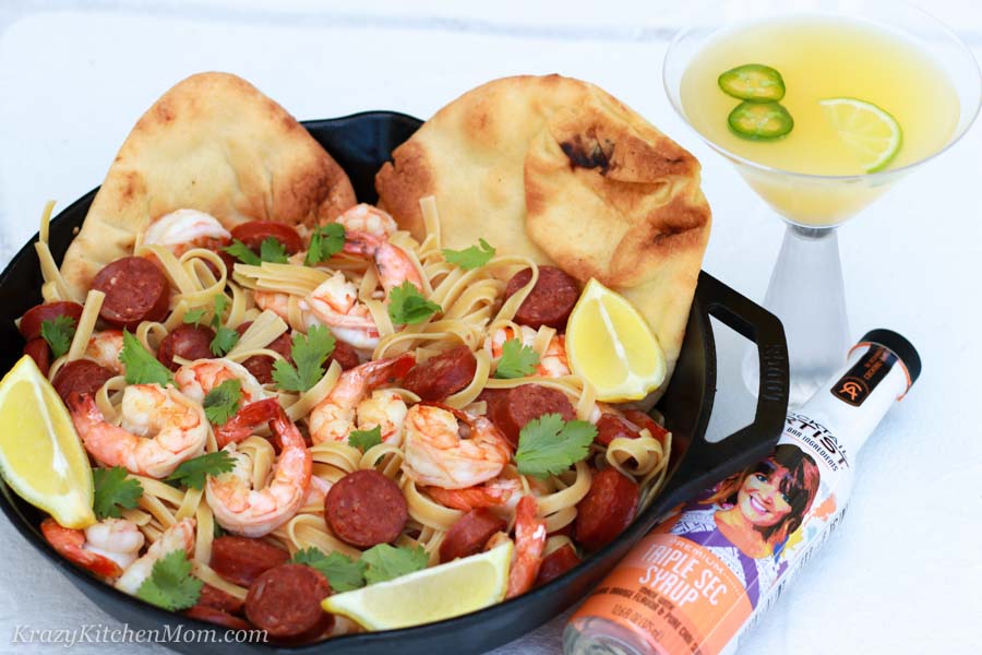 a pan of pasta, shrimp and sausage with a slice of lime and two pitas and a cocktail