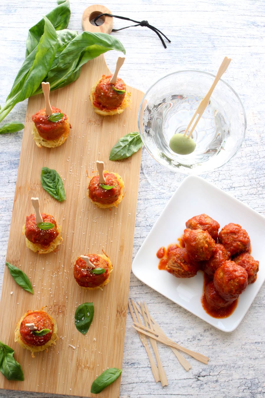 six mini meatball skewers on a board with a side of meatballs and a martini with an olive on a stick