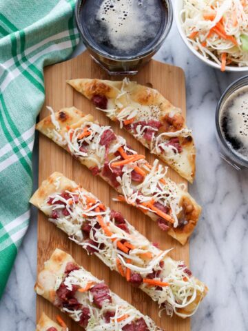 Corned beef flatbread on a wood board, with a bowl of slaw and two dark beers