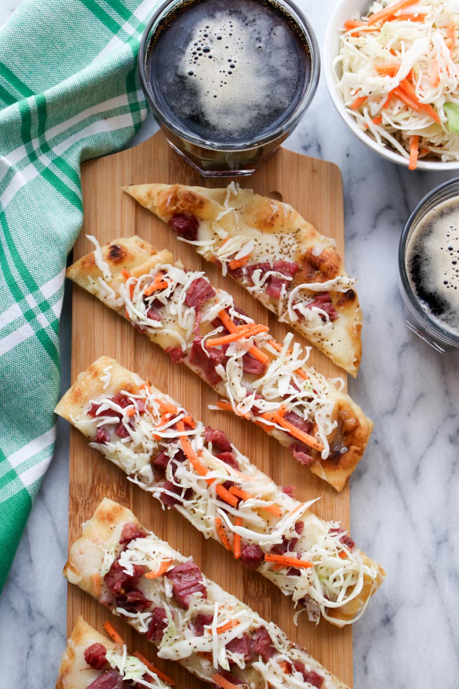 Corned beef flatbread on a wood board, with a bowl of slaw and two dark beers