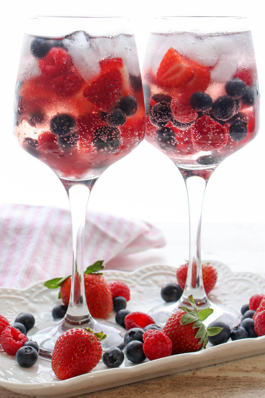 TWO GLASSES OF PINK BERRY GIN AND TONIC ON A WHITE TRAY WITH BERRIES SCATTERED ABOUT