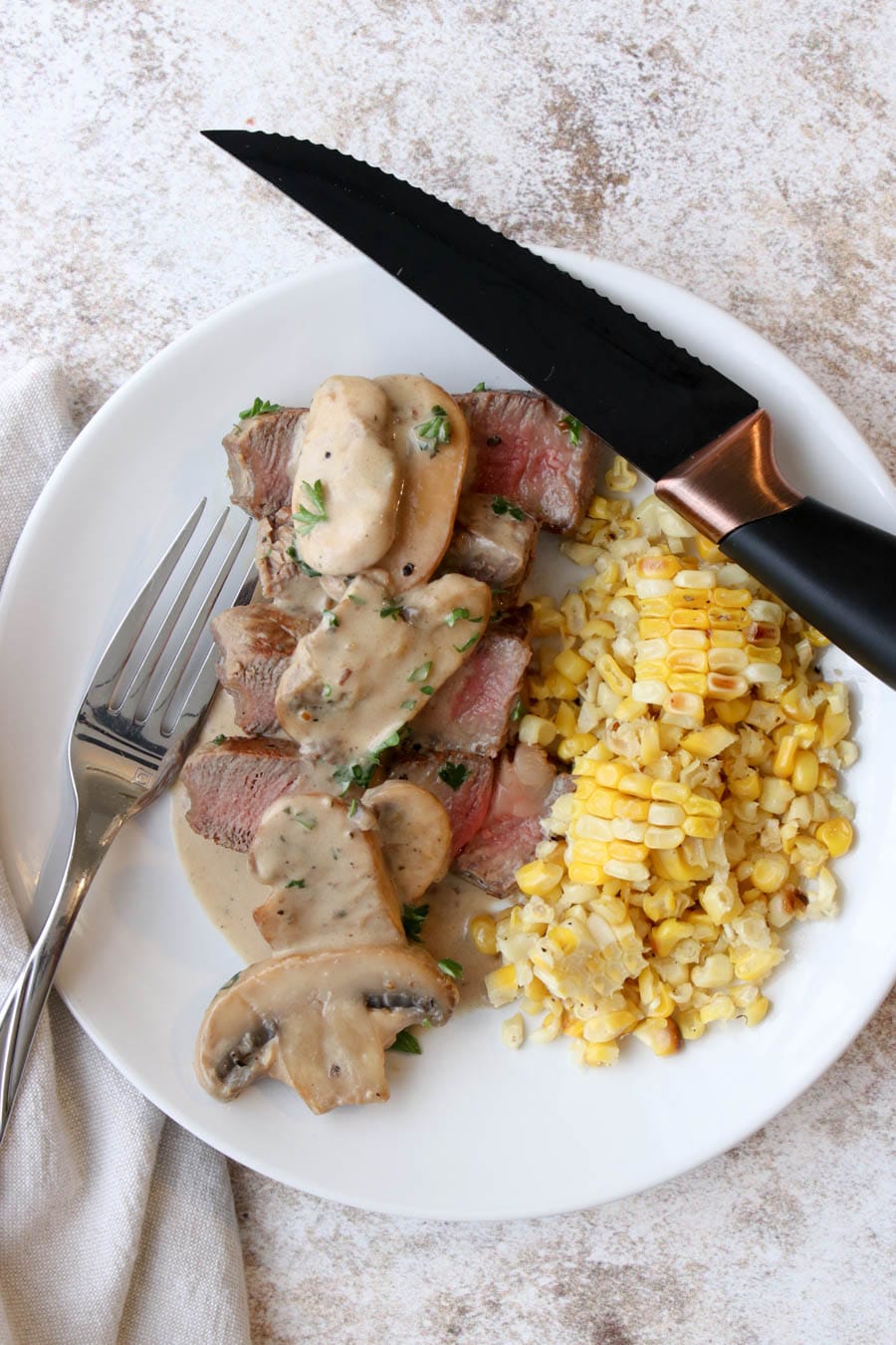 a white plate with slices of steak covered with a mushroom cream sauce and corn on the side.