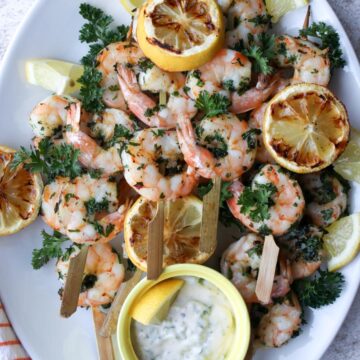 white platter with shrimp skewers and grill lemons on it with a side of white dipping sauce