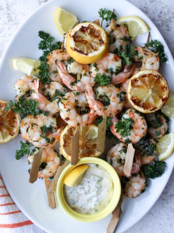 white platter with shrimp skewers and grill lemons on it with a side of white dipping sauce