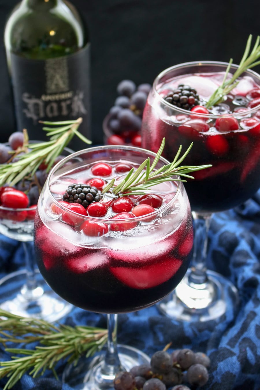 Two wine glasses filled with red wine and a rosemary sprig with bottle of wine in background