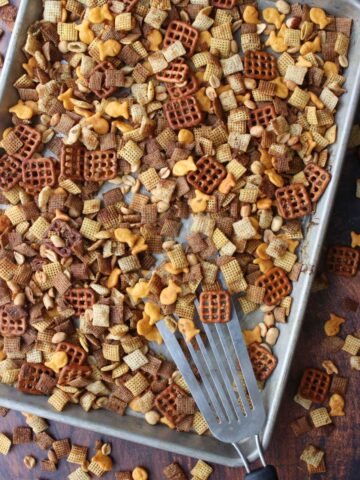 Chex party mix on a cookies sheet being scooped with a metal spatula