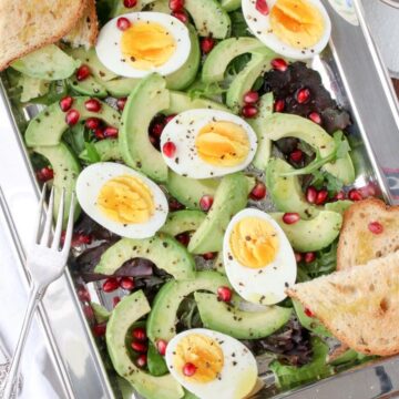 silver platter filled with lettuce, avocados, hard boiled eggs, pomegranate seeds and toast