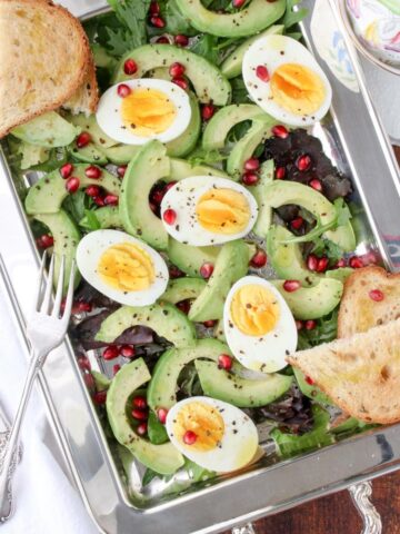 silver platter filled with lettuce, avocados, hard boiled eggs, pomegranate seeds and toast
