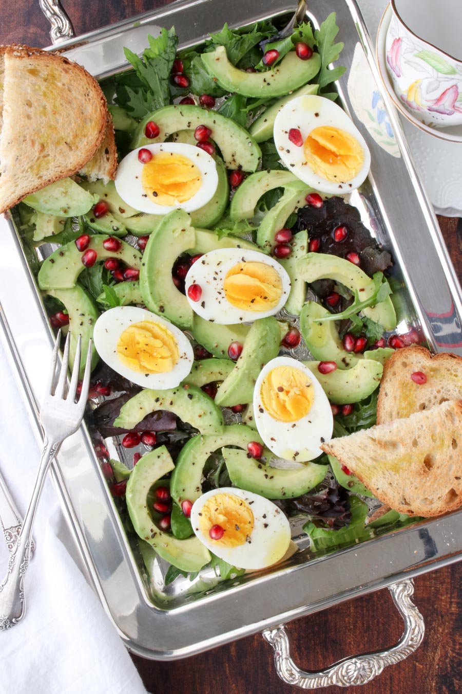 silver platter filled with lettuce, avocados, hard boiled eggs, pomegranate seeds and toast