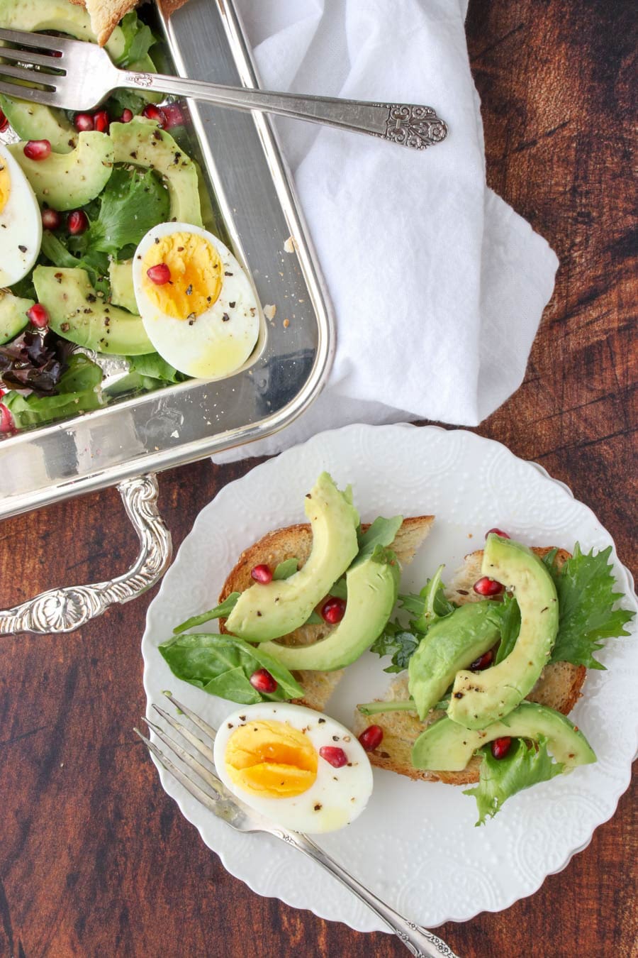 while plate with two slices of toast with sliced avocado on top and hard-boiled egg on the side, garnished with pomegranate seeds