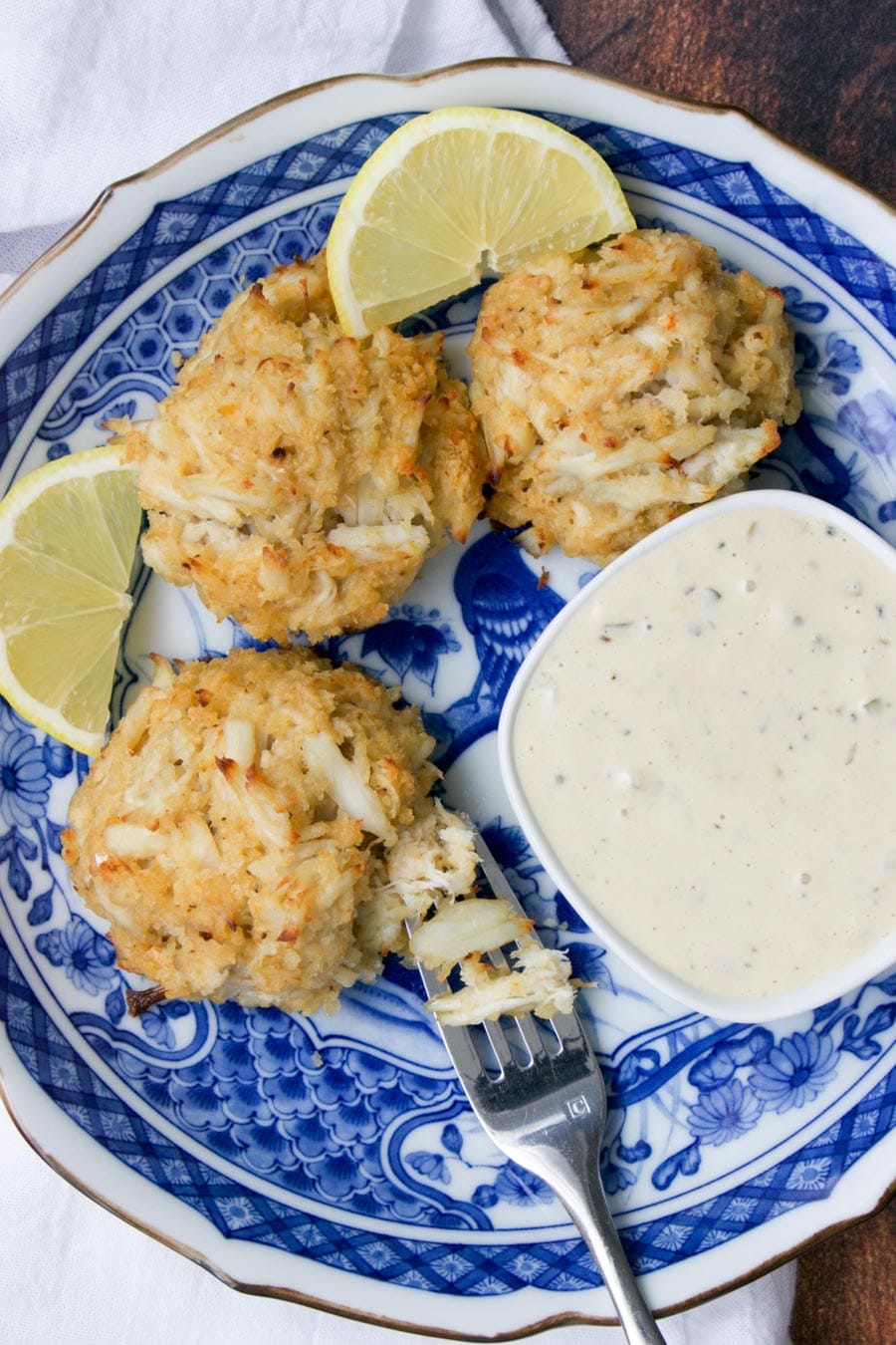 three crab cakes, bowl of sauce, and lemon slices on a blue plate