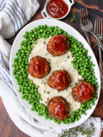 platter of mashed potatoes, surrounded by green peas and topped with mini meatloaf meatballs