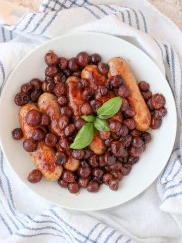 a white plate with cooked sausages, grapes and a sprig of fresh basil