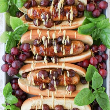 a tray with four sausages in buns topped with grapes and fresh basil