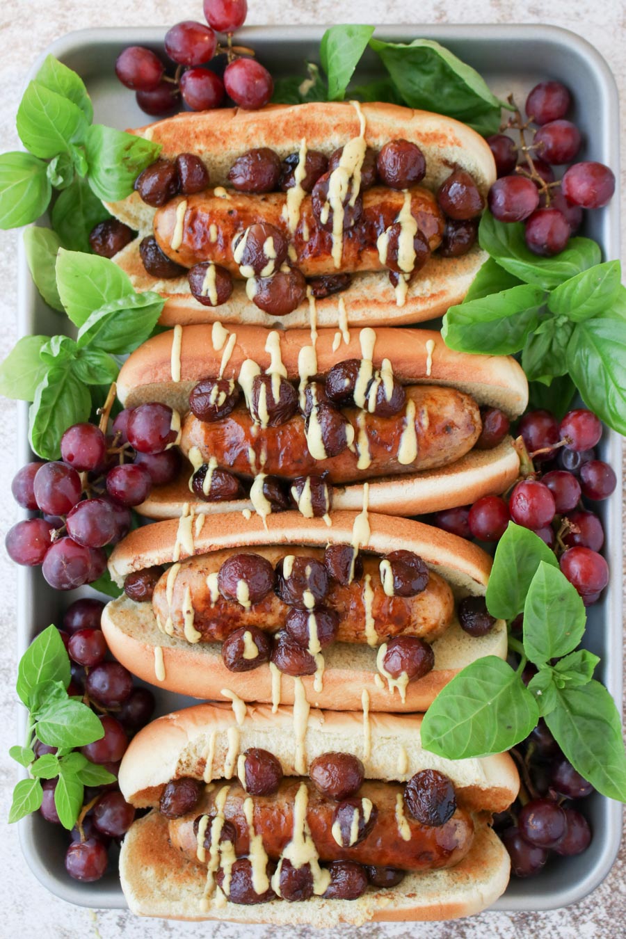 a tray with four sausages in buns topped with grapes and fresh basil