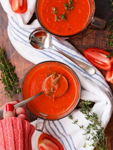 Ladies hand with a spoon of tomato soup next to another bowl of soup and a blue and white cloth