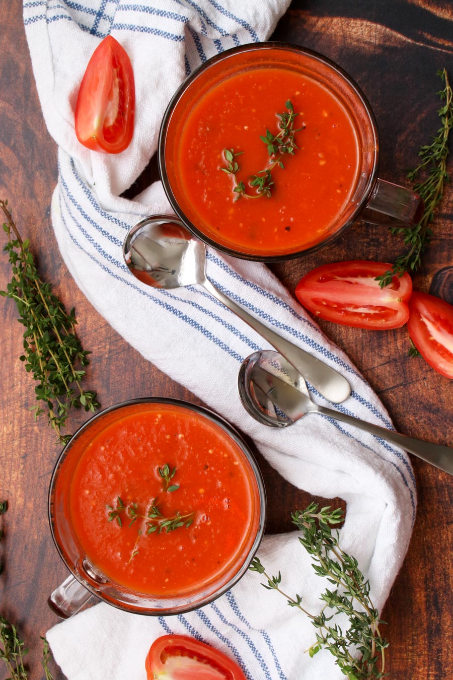 two bowls of tomato soup with a blue and white cloth around them, two spoons and some thyme and sliced tomatoes