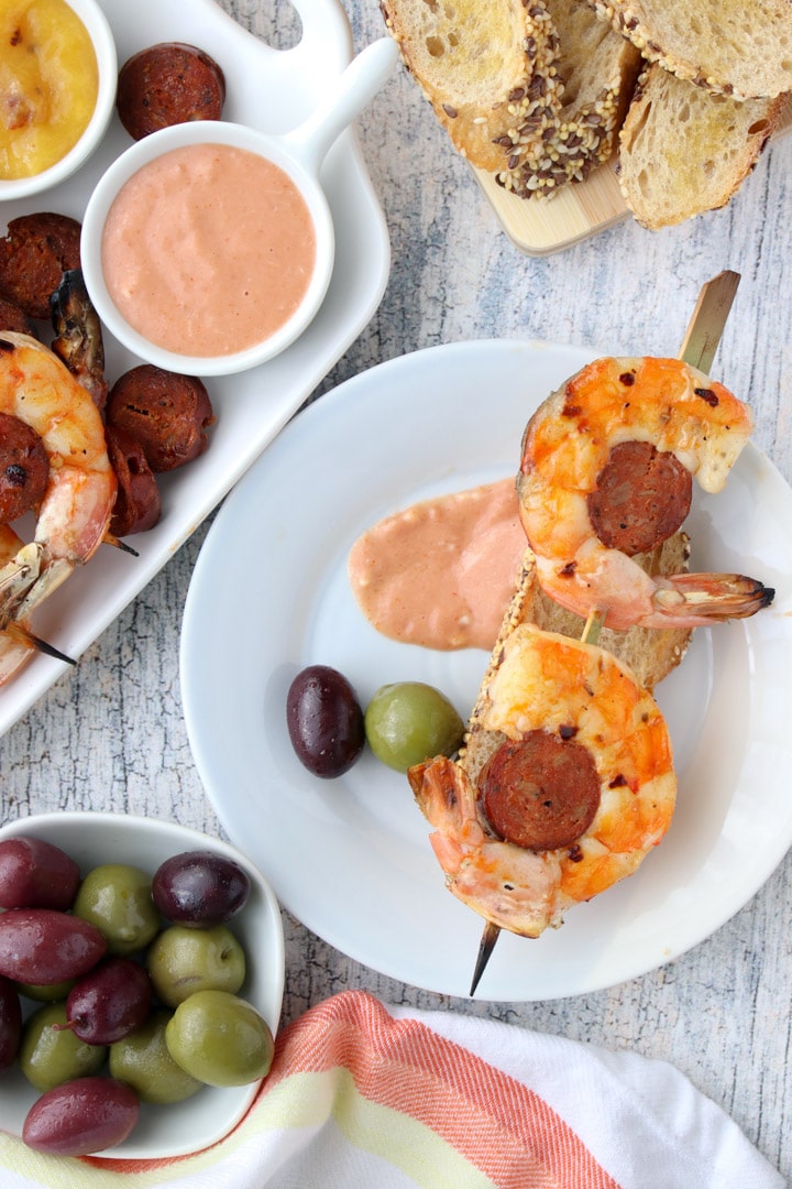 small plate with a shrimp and chorizo skewer, two olives and a slice of toasted bread