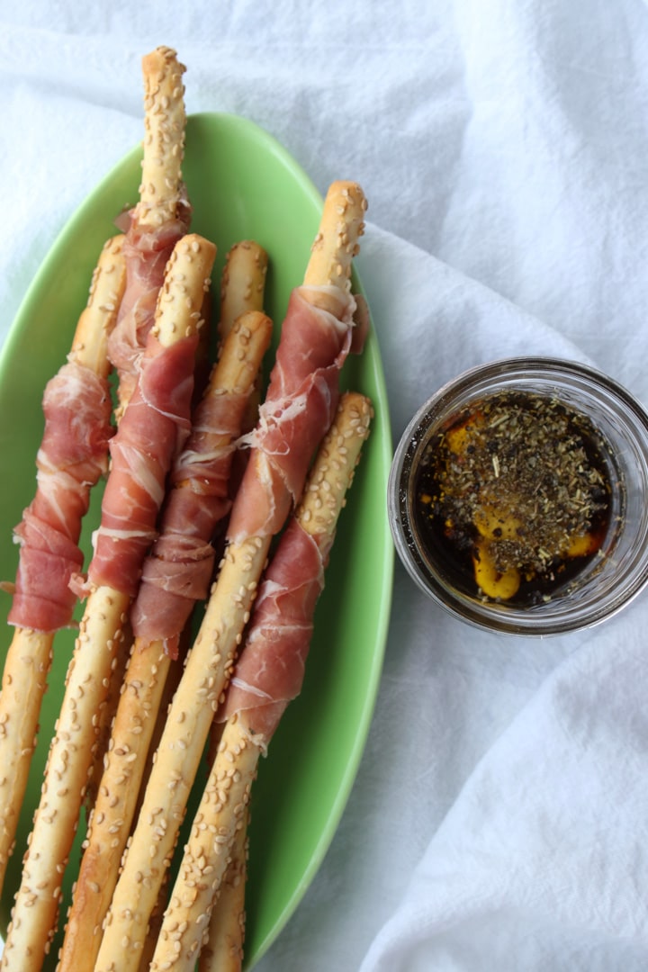 prosciutto wrapped breadsticks on a green plate with a bowl of vinaigrette on the side 