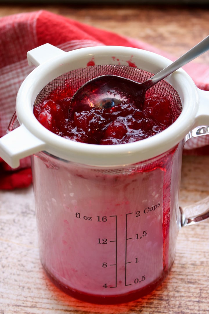 cooked cranberries being strained into a glass measuring cup