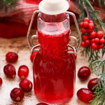 BOTTLE OF CRANBERRY SYRUP WITH FRESH CRANBERRIES AND WINTER GREENS IN BACKGROUND