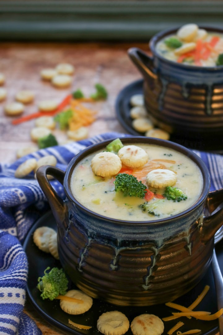 two blue and brown soup bowls with broccoli cheese soup topped with crackers, broccoli, carrots