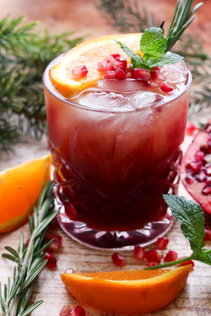 Pomegranate vodka cocktail topped with fresh orange slice, pomegranate arils, fresh mint and rosemary