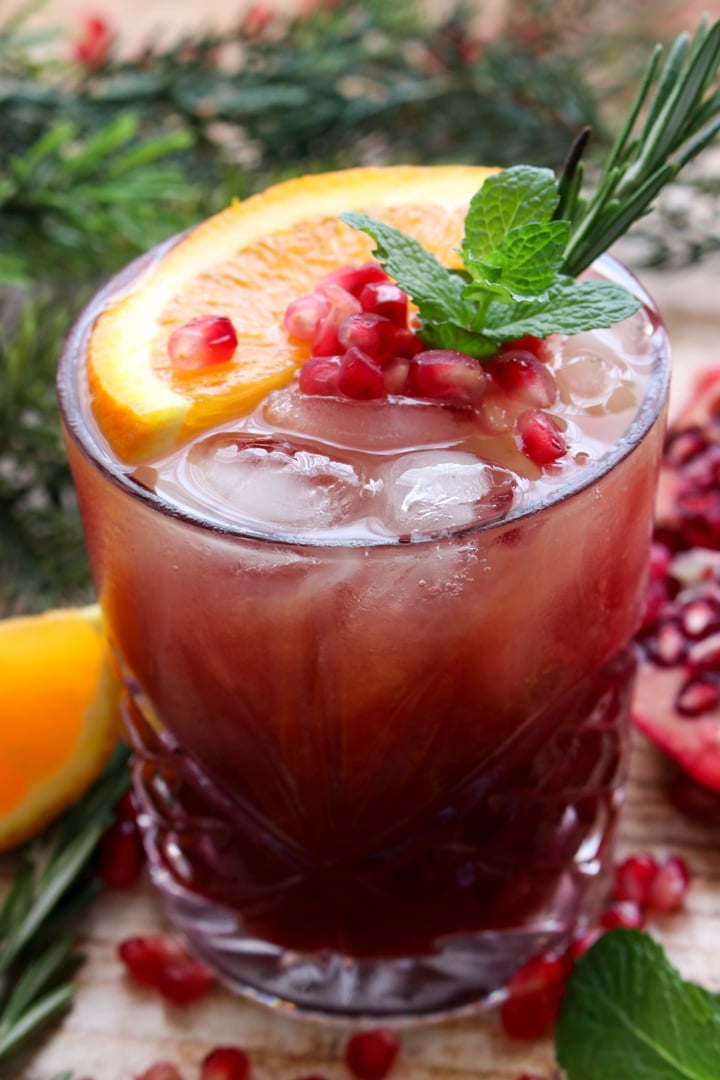 close up shot of pomegranate cocktail garnished with an orange slice, pomegranate seeds, rosemary and mint