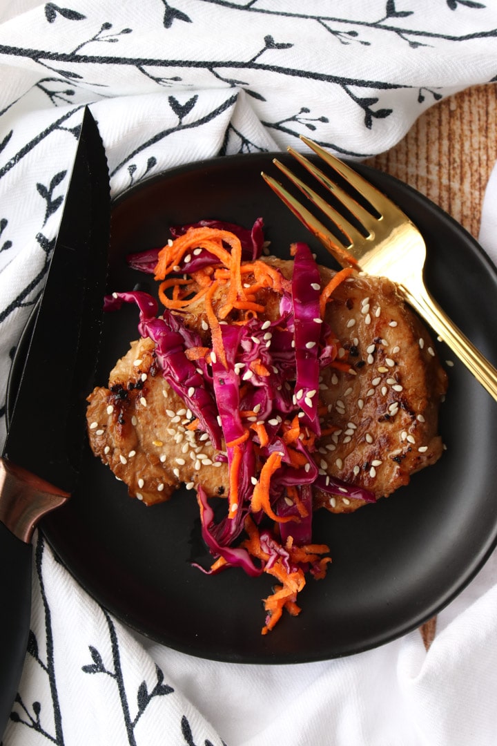 overhead shop of pork chop on a black plate with red cabbage carrot slaw on top with fork and knife