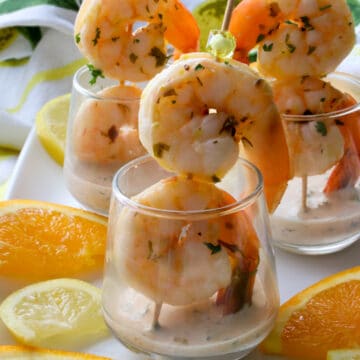 two shrimp on a skewer in a glass with citrus sauce and lemon and oranges slices on the side