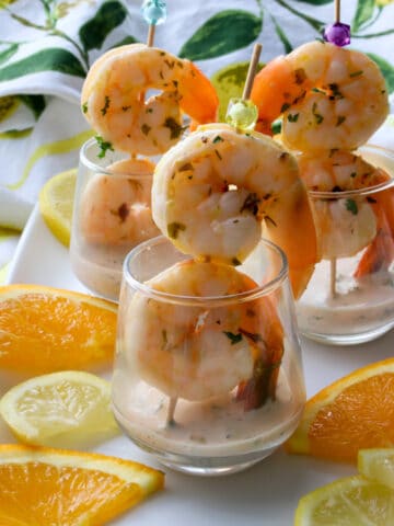 two shrimp on a skewer in a glass with citrus sauce and lemon and oranges slices on the side