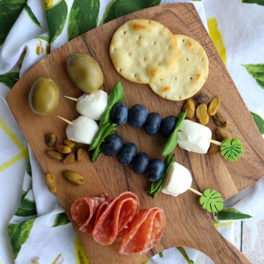 two blueberry skewers on a small board with olives, crackers, and salami