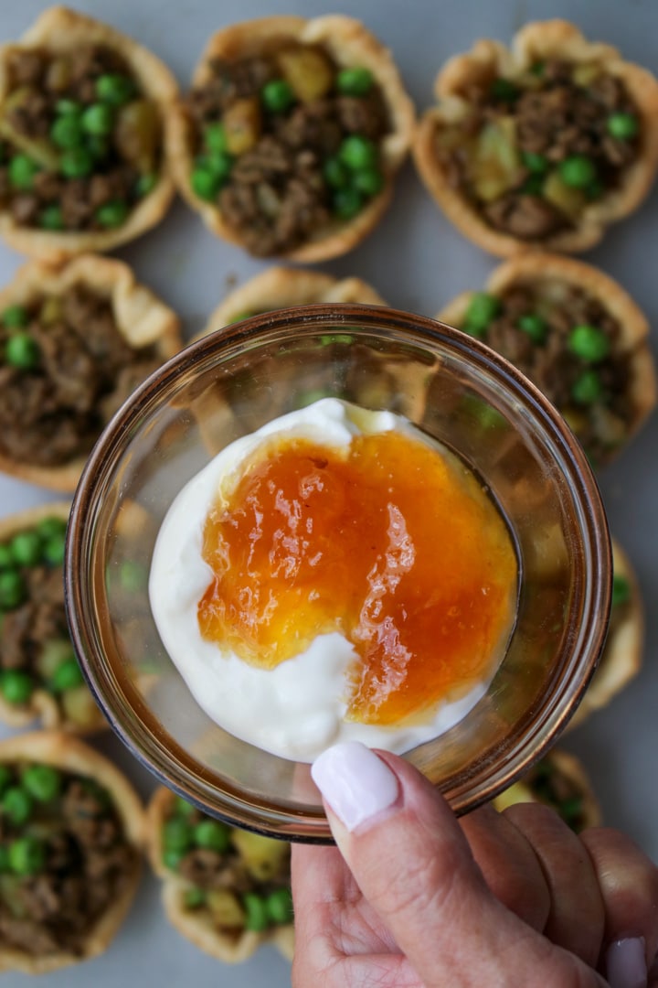 woman's hand holding a bowl of greek yogurt and mango chutney over a tray of curried beef pies