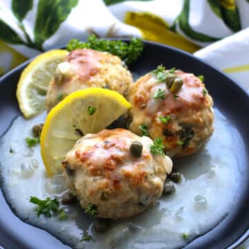 Three lemon caper chicen meatballs on a black plate with lemon slices and lemon sauce