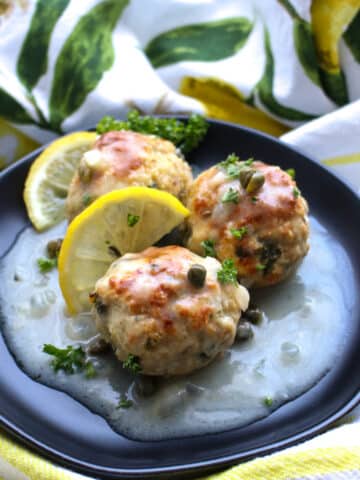 Three lemon caper chicen meatballs on a black plate with lemon slices and lemon sauce