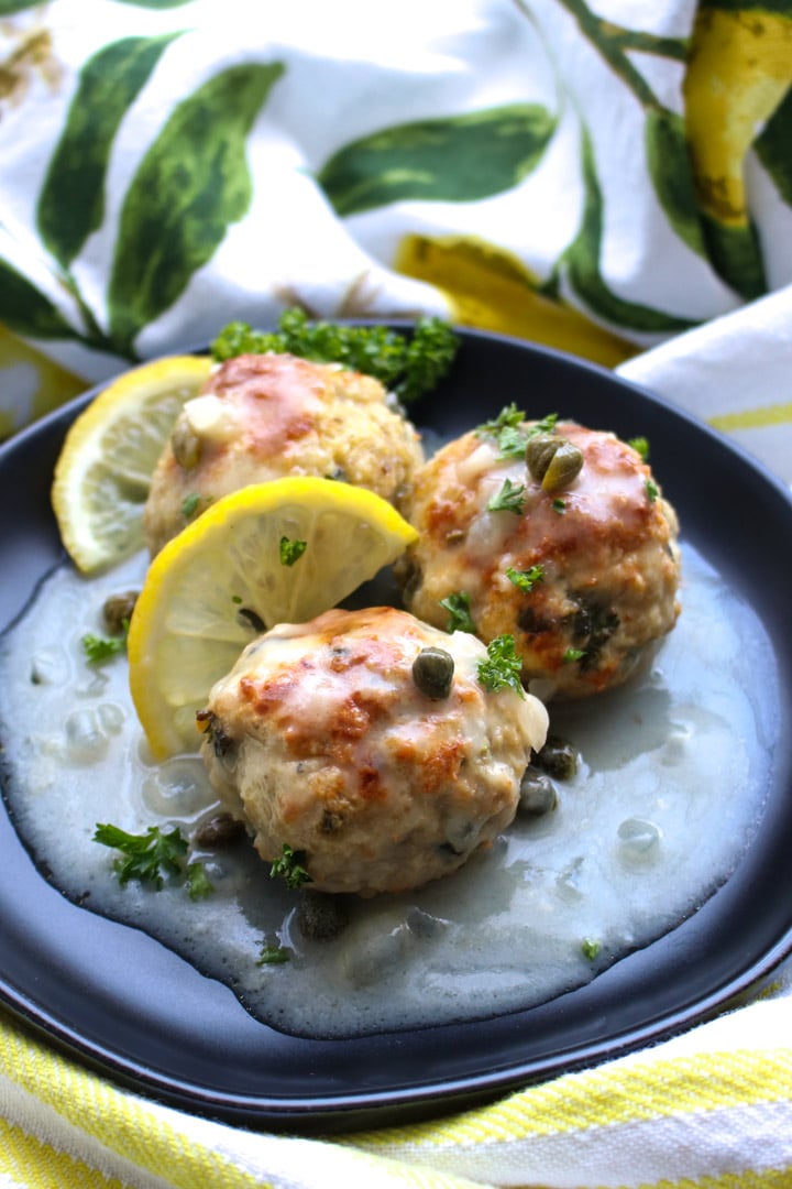 Three lemon caper chicen meatballs on a black plate with lemon slices and lemon sauce